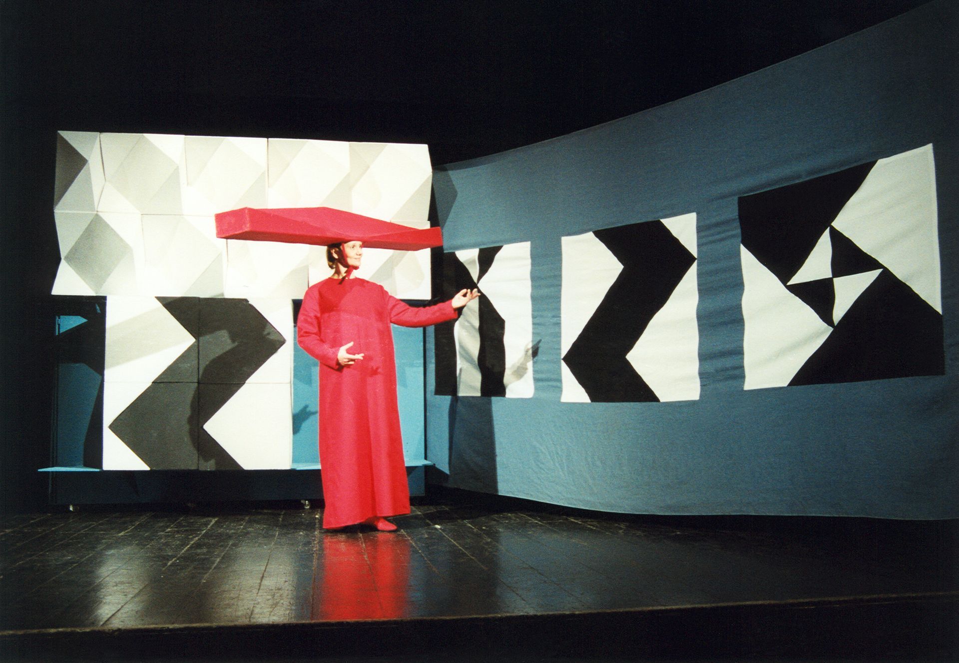 Aktorka w czerwonym kostiumie i kapeluszu w formie podłużnej bryły.