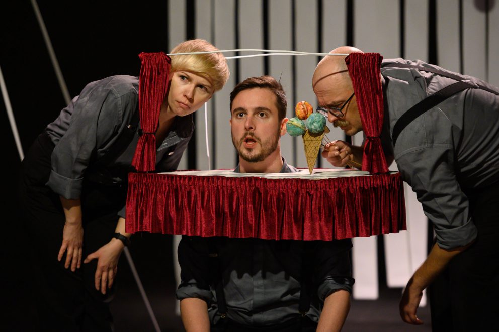 Trójka aktorów z głowami w mini scenie. Jeden z nich trzyma loda z trzema kolorowymi gałkami.