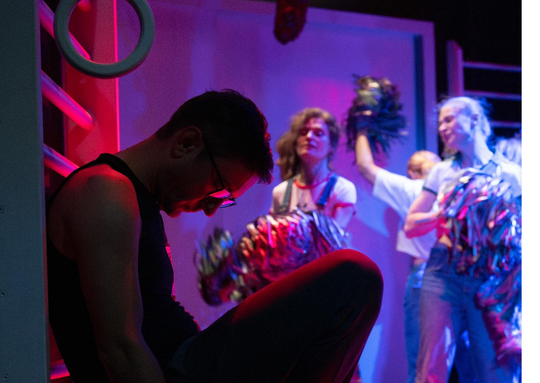 Chłopak w okularach siedzi ze spuszczoną głową i opiera się o konstrukcję w czerwono-fioletowym oświetleniu. W tle widać trzy osoby tańczące lub wykonujące ruchy z kolorowymi pomponami.